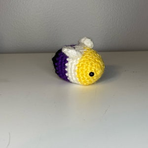 May include: A crocheted bee with purple, white, and yellow stripes. The bee has a small black eye and a white wing.