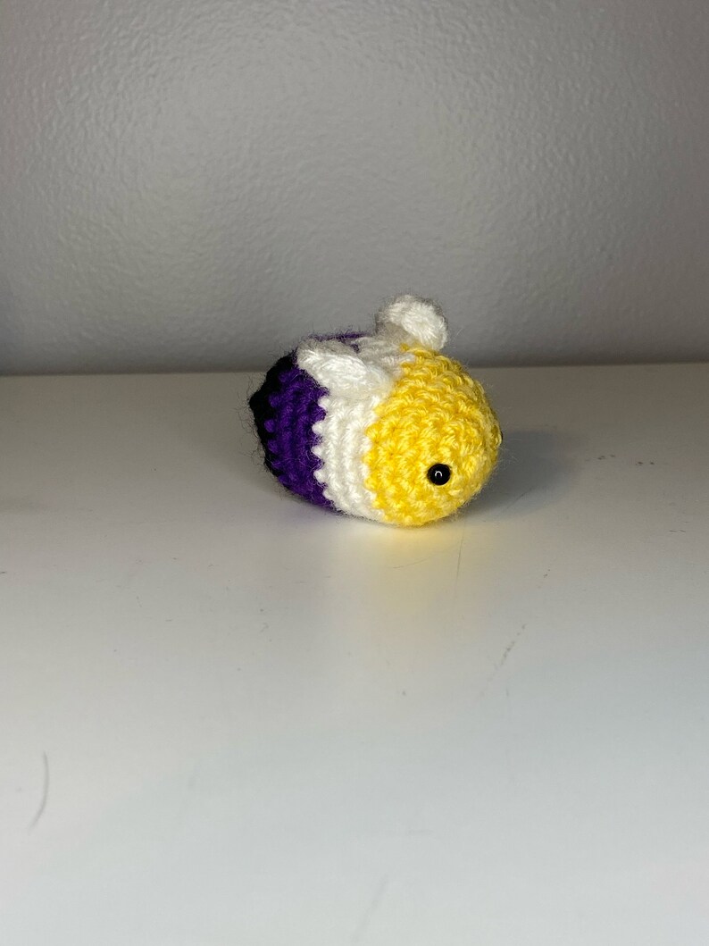May include: A crocheted bee with purple, white, and yellow stripes. The bee has a small black eye and a white wing.