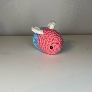 May include: A crocheted bee toy with pink and blue stripes and white wings. The bee has black eyes.