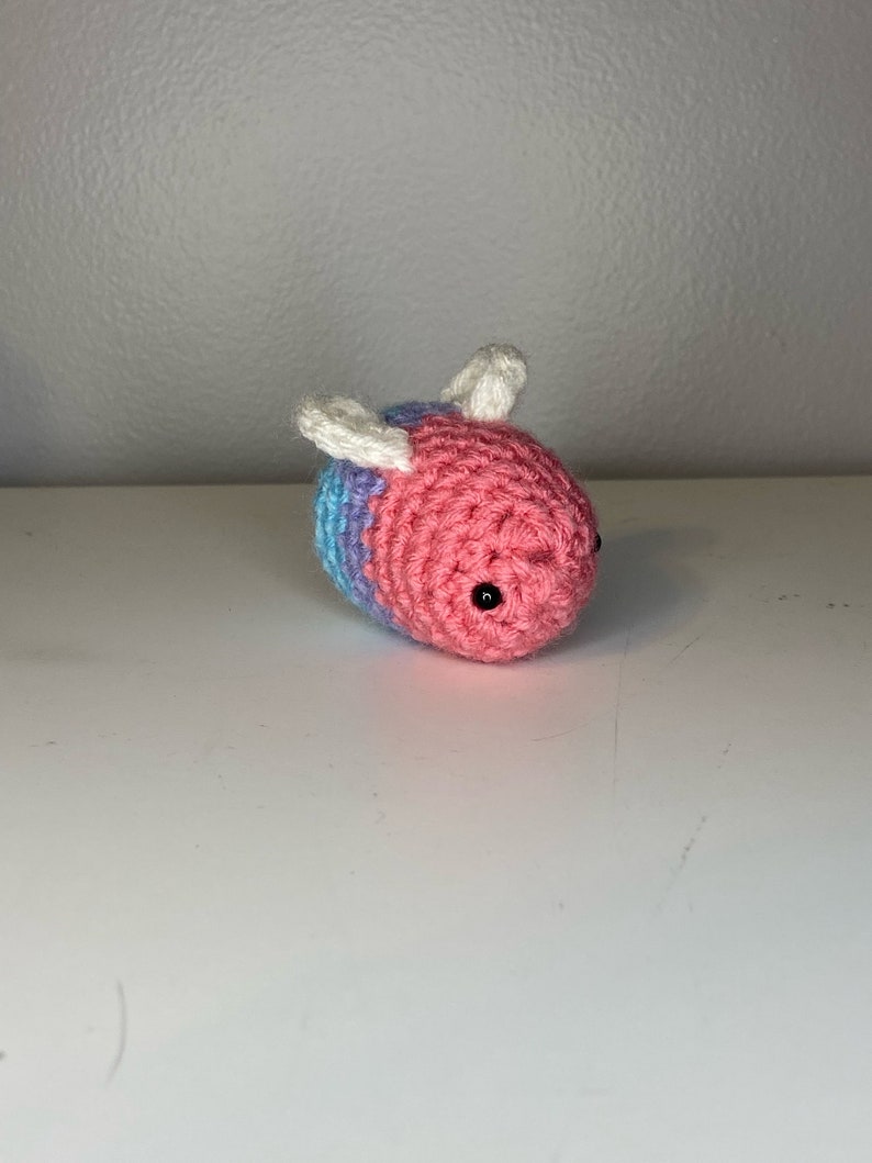 May include: A crocheted bee toy with pink and blue stripes and white wings. The bee has black eyes.