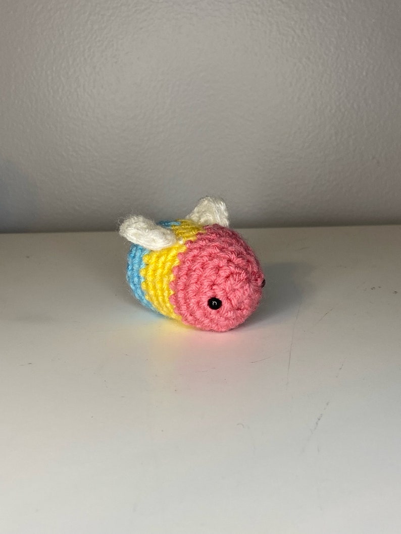 May include: A crocheted bee toy with blue, yellow, and pink stripes. The bee has black eyes and white wings.