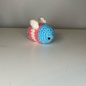 May include: A crocheted bee toy with blue, pink, and white stripes. The bee has white wings and black eyes.