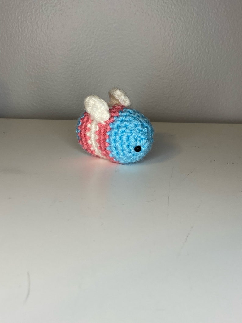 May include: A crocheted bee toy with blue, pink, and white stripes. The bee has white wings and black eyes.
