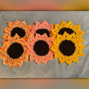May include: Four crocheted sunflower dishcloths in shades of orange, yellow, and pink. The dishcloths have a brown center and a crocheted border.