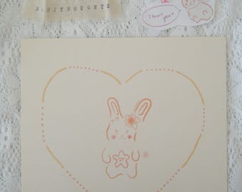 May include: A square off-white card featuring a light orange line drawing of a rabbit inside a heart shape. The rabbit has a flower on its head and holds a star. Above the card are stickers including a rabbit with the text I heart you.