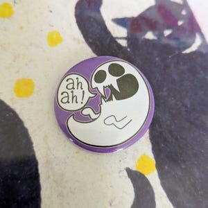 May include: A round, purple button with a cartoon ghost design. The ghost is white with a skull-like face and is saying "ah ah!" in a speech bubble. The background has a cat silhouette and yellow dots.