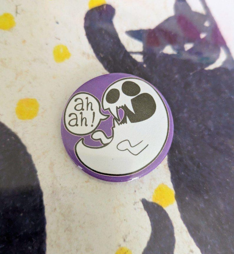 May include: A round, purple button with a cartoon ghost design. The ghost is white with a skull-like face and is saying "ah ah!" in a speech bubble. The background has a cat silhouette and yellow dots.