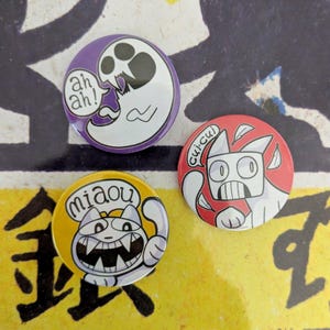 May include: Three round pinback buttons with cartoon cat and ghost designs. One button is purple with a white ghost and the text "ah ah!". Another is red with a white cat and the text "cui-cui". The last is yellow with a white cat and the text "miaou".