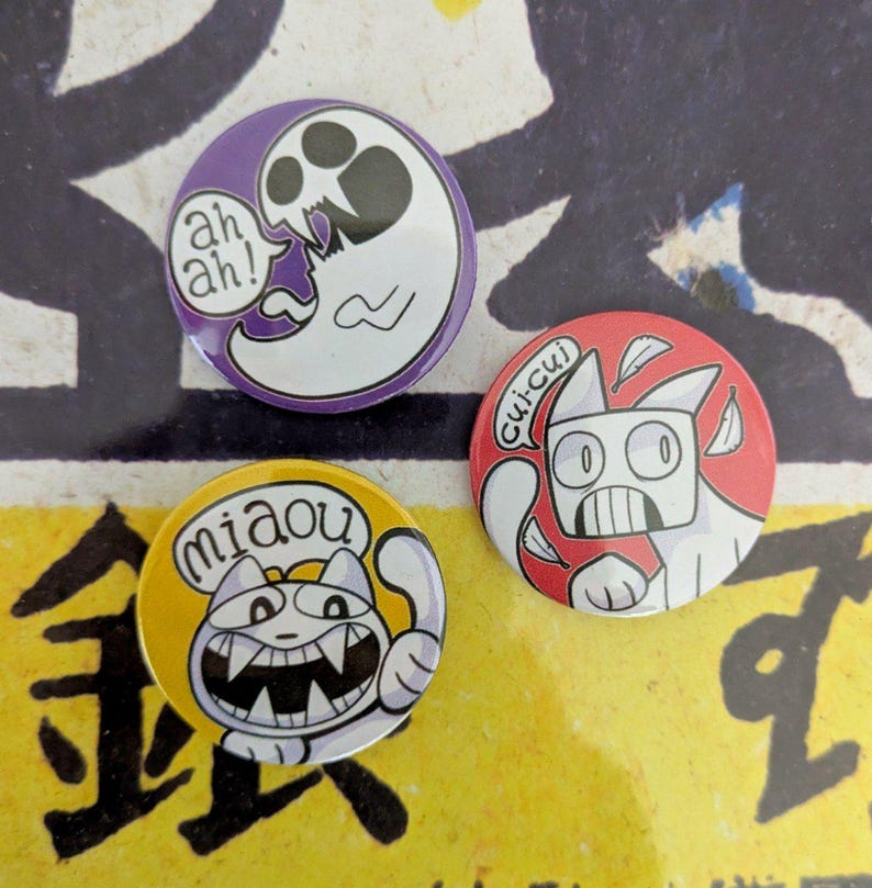 May include: Three round pinback buttons with cartoon cat and ghost designs. One button is purple with a white ghost and the text "ah ah!". Another is red with a white cat and the text "cui-cui". The last is yellow with a white cat and the text "miaou".
