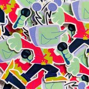 Op de afbeelding: Een verzameling kleurrijke stickers met een stripfiguur met een tv-hoofd, groene huid en een rode jas. Het personage houdt een microfoon vast en heeft een geel bliksemontwerp op de jas. De stickers hebben een retro, pop-art stijl.