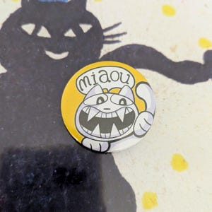 May include: A round, yellow button with a cartoon cat illustration. The cat has a wide grin, sharp teeth, and is giving a thumbs up. The word "miaou" is in a speech bubble. A black cat silhouette is in the background.