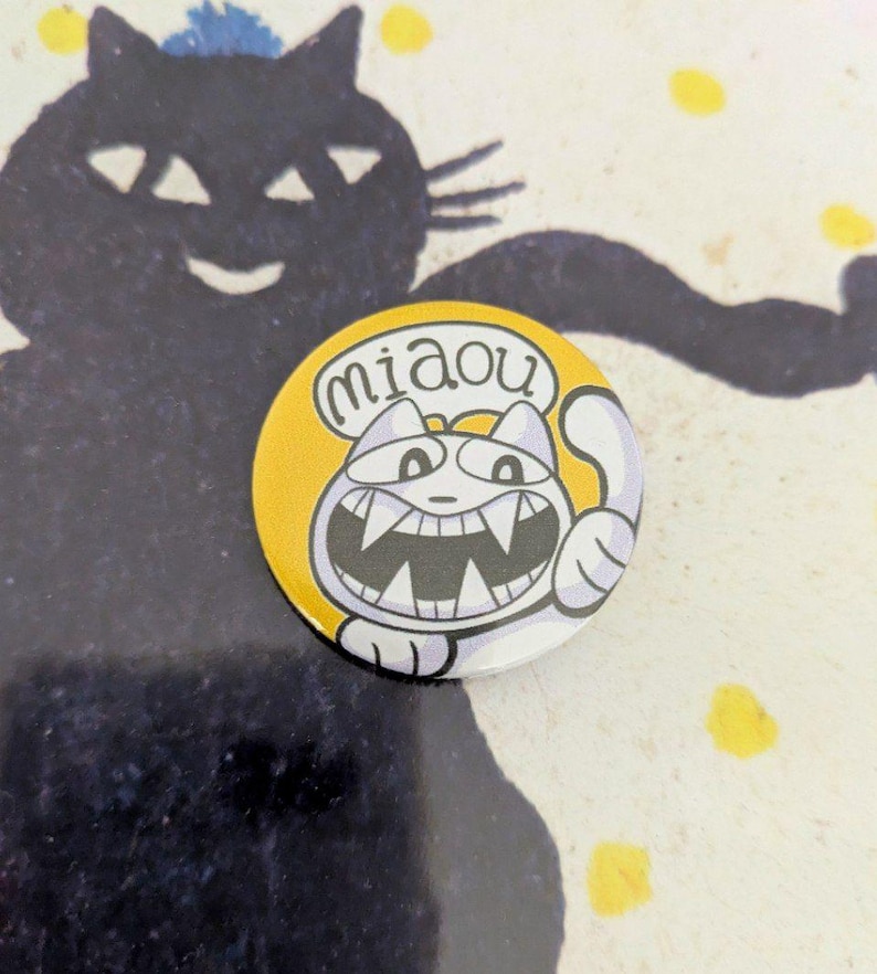 May include: A round, yellow button with a cartoon cat illustration. The cat has a wide grin, sharp teeth, and is giving a thumbs up. The word "miaou" is in a speech bubble. A black cat silhouette is in the background.