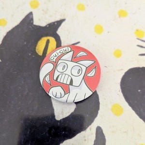 May include: A round, red and white button featuring a cartoon cat with the text "Cui-cui" in a speech bubble. The cat has a surprised expression and is surrounded by white leaf shapes. The background is a patterned surface.