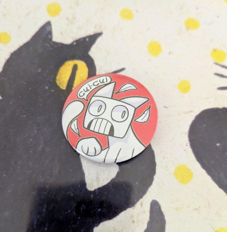 May include: A round, red and white button featuring a cartoon cat with the text "Cui-cui" in a speech bubble. The cat has a surprised expression and is surrounded by white leaf shapes. The background is a patterned surface.