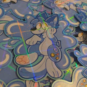 May include: A collection of holographic stickers featuring a cartoon character with blue and white fur, holding a palette with a smiley face, star, and other shapes. The stickers have a unique, wavy outline.