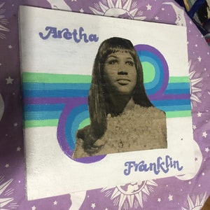 May include: A white canvas painting with a black and white photo of Aretha Franklin. The painting has a purple, blue, and green striped design behind the photo. The text "Aretha Franklin" is written in purple cursive.