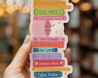 May include: A vertical sticker featuring a stack of colorful books with romance tropes printed on the spines, including Love at First Sight, Enemies to Lovers, Marriage of Convenience, Soul Mates, Forbidden Love, Love Triangle, Fake Dating, Age Gap, One Bed, Sloooow Burn, Forced Proximity, Fated Mates, and Second Chance, with a pink mug and white daisies.