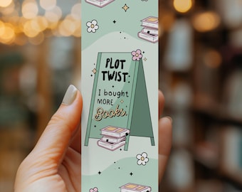 May include: A rectangular bookmark with a light green background featuring illustrations of stacked books, small white flowers, and stars. The center displays a green A-frame sign with the text: PLOT TWIST: I bought more Books.