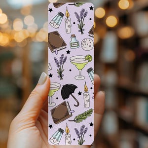 May include: A rectangular bookmark with a light purple background featuring a repeating pattern of brown books, lit candles, lavender sprigs, salt shakers, small glass bottles, a black umbrella, a moon, and a cocktail glass with a lime slice.