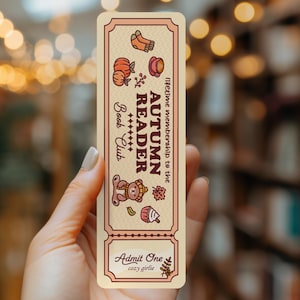 May include: A rectangular bookmark with a cream background and brown border, featuring the text "AUTUMN READER Book Club" and "lifetime membership to the", with illustrations of pumpkins, a cup, a bear, a leaf, and "Admit One cozy girlie".