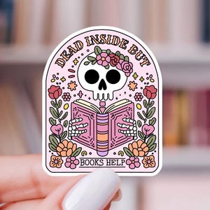 May include: A die-cut sticker featuring a white skull wearing a pink floral crown while reading an open pink book. The text reads DEAD INSIDE BUT BOOKS HELP. The design includes colorful flowers, green leaves, and small yellow stars on a light pink background.