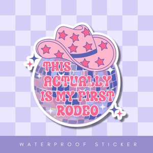 May include: A pink and blue disco ball sticker with a pink cowboy hat on top. The sticker says "This Actually Is My First Rodeo".