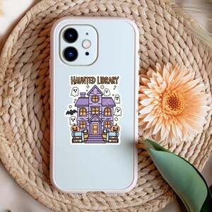May include: A white phone case featuring a purple haunted library design. The design includes a spooky house with ghosts, bats, and pumpkins. The text "Haunted Library" is printed on the case.