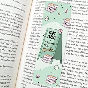 May include: A green bookmark with a white border and a stack of books on a stand. The bookmark has the text "Plot Twist: I bought more books" in white letters. The background is a light green with white stars and flowers.