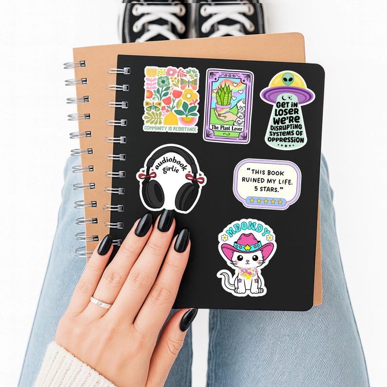 May include: A black spiral-bound notebook decorated with six stickers: a floral arrangement with the text "COMMUNITY IS RESISTANCE," a tarot card labeled "The Plant Lover," a UFO with "GET IN LOSER WE'RE DISRUPTING SYSTEMS OF OPPRESSION," headphones with "audiobook girlie," a five-star review bubble, and a cat in a pink cowboy hat with "MEOWDY."