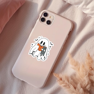 Peut inclure: Une coque de t&eacute;l&eacute;phone rose avec un autocollant blanc repr&eacute;sentant un fant&ocirc;me lisant un livre avec un chat noir. Le fant&ocirc;me est blanc et a un visage amical. Le livre est orange et a une reliure noire. Le chat est noir et a un visage blanc. L'autocollant est sur une coque de t&eacute;l&eacute;phone rose.
