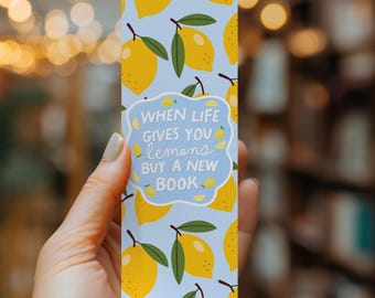 May include: A rectangular bookmark with a light blue background featuring a repeating pattern of yellow lemons with green leaves. In the center, a white scalloped label displays the text: When life gives you lemons buy a new book.