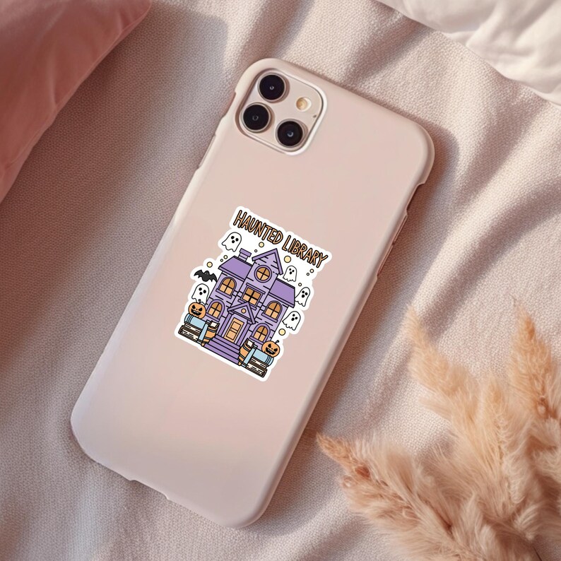 May include: A pink phone case with a sticker of a purple haunted library building with ghosts and pumpkins. The sticker says "Haunted Library".