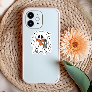 Peut inclure: Un fant&ocirc;me blanc tenant un livre et un chat noir dans ses bras. Le fant&ocirc;me est entour&eacute; d'&eacute;toiles et de chauves-souris. L'image est sur une coque de t&eacute;l&eacute;phone blanche.