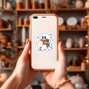 Peut inclure: Un smartphone blanc avec un autocollant repr&eacute;sentant un fant&ocirc;me tenant un livre et un chat sur l'&eacute;cran.