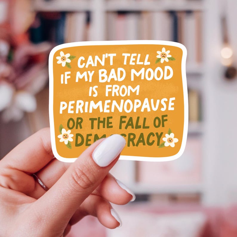 May include: A yellow sticker with a white border and a humorous message about perimenopause and the state of democracy. The sticker features a daisy with a sad face on each side of the text. The text reads: "Can't tell if my bad mood is from perimenopause or the fall of democracy."