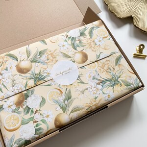 May include: A cardboard box with a yellow and white floral pattern wrapping paper. The paper features lemons, oranges, and white flowers. A white circle sticker with the text "handgemacht f&uuml;r dich" is on the box.