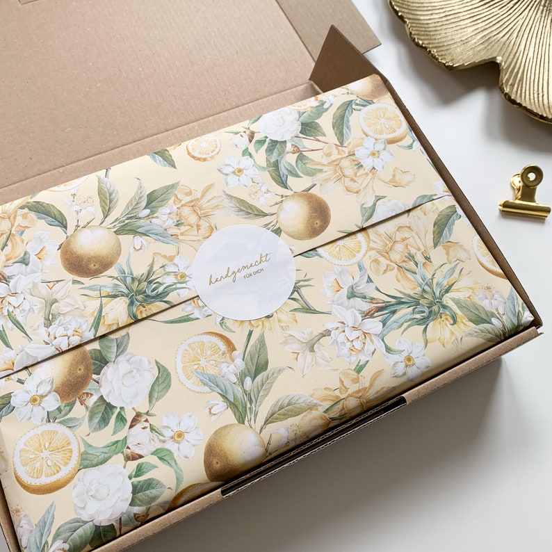 May include: A cardboard box with a yellow and white floral pattern wrapping paper. The paper features lemons, oranges, and white flowers. A white circle sticker with the text "handgemacht f&uuml;r dich" is on the box.