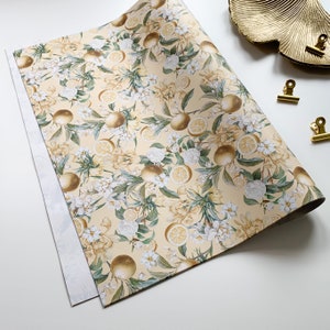 May include: A sheet of yellow wrapping paper with a floral pattern featuring white flowers and lemons. The paper has a subtle texture and a slightly glossy finish.