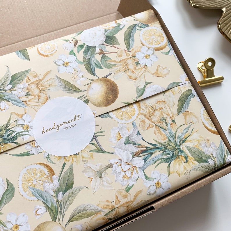May include: A light brown box with a floral wrapping paper design. The paper features white flowers, green leaves, and yellow lemons. A white circle sticker with the text "handgemacht f&uuml;r dich" is on the box.