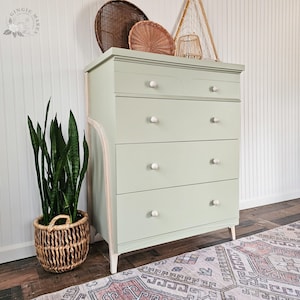 May include: A sage green chest of drawers with four large drawers and two smaller top drawers. The chest has white knobs and a light wood trim. A woven basket with a tall green plant sits to the left. Decorative woven items are on top.