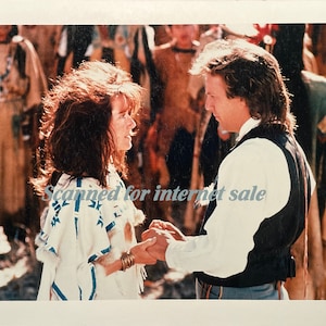 May include: A wedding ceremony with a couple in traditional clothing. The woman is wearing a white dress with blue and white patterns. The man is wearing a white shirt and a black vest. The ceremony is taking place outdoors with a group of people in the background.