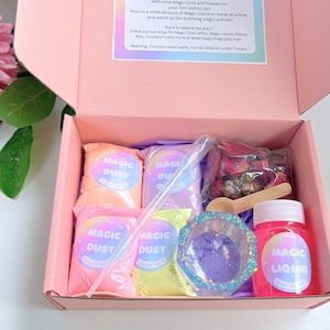 May include: A pink box filled with a magic potion kit. The kit includes bags of colorful "Magic Dust", a small bottle of "Magic Liquid", a clear dropper, a wooden spoon, and a decorative container. The box also contains dried flowers.