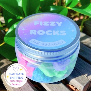 May include: A clear plastic jar with a silver screw-top lid containing colourful, irregular-shaped fizzy rocks. The label on the lid features a holographic gradient background with the white text Happy Magic Play FIZZY ROCKS Just add water.