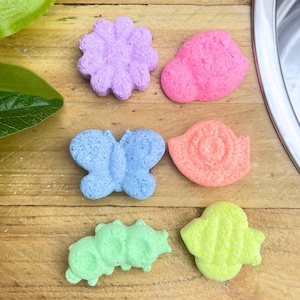 May include: Six colourful bath bombs in various shapes including a purple flower, a pink cloud, a blue butterfly, an orange snail, a green caterpillar, and a yellow bee. Each piece has a textured, porous surface and is arranged on a wooden surface.