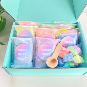 May include: A light blue box containing packets of colourful "Magic Dust" and "Fizzy Salts", plus a small wooden scoop. The packets are in pink, orange, blue, and green. Some packets have the text "Just add water".
