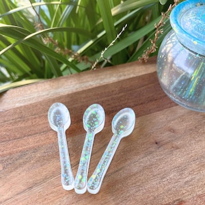 May include: Three clear resin spoons with iridescent glitter. The spoons are arranged on a wooden surface. A jar with a blue lid and glitter is in the background. Green foliage is visible.