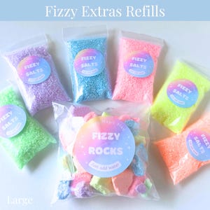 May include: Assortment of colorful bath product refills in clear plastic bags. The bags contain fizzy salts and rocks in various colors, including purple, blue, pink, green, and orange. Each bag has a round label that reads "Fizzy Salts" or "Fizzy Rocks". The text "Just add water" is also visible.