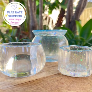 May include: Three clear resin containers with iridescent glitter accents. The set includes a rounded bowl, a wider jar with a flared rim, and a smaller cylindrical vessel. Each piece features a smooth, glossy finish with embedded colourful sparkles.