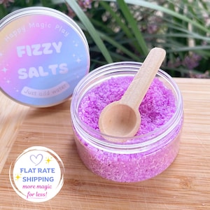 May include: A clear plastic jar filled with vibrant purple fizzy salts and a small wooden scoop. The jar lid is nearby, featuring a holographic label with the text Happy Magic Play, FIZZY SALTS, and Just add water, decorated with small white stars.