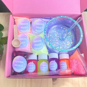 May include: A pink box containing a DIY slime kit. The kit includes bags of "Magic Dust", bottles of "Magic Liquid", a bowl with glitter, a wooden spoon, and a bag of "Potion Petals". The kit is designed for making slime.